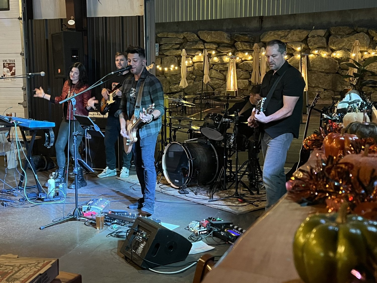 A live band performing indoors. The band includes a woman singing, a male guitarist, and a drummer in the background, with a stone wall and string lights behind them.