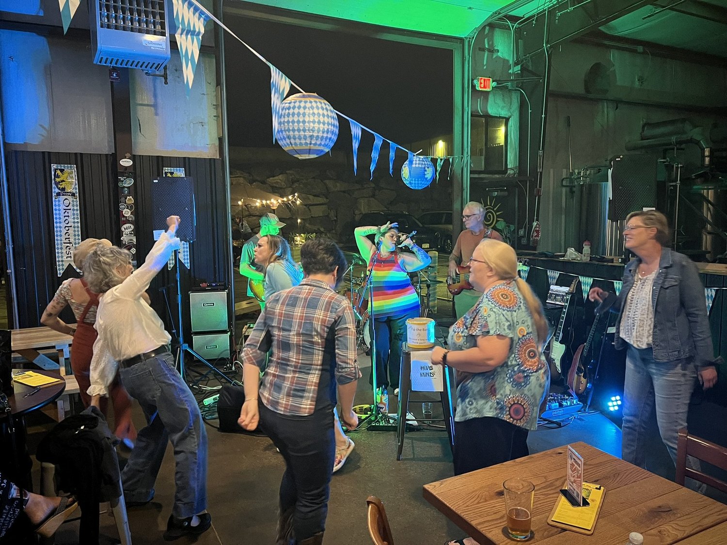 A lively indoor party scene with a live band performing on stage, decorated with blue and white bunting and paper lanterns. People are dancing and socializing around tables, with drinks in hand, and colorful lighting creating a festive atmosphere.