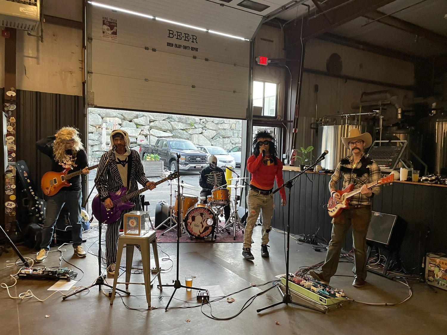 A band of five musicians performing indoors with guitars and drums, dressed in various costumes and sunglasses, in a rustic setting with an open garage door showing parked cars outside.