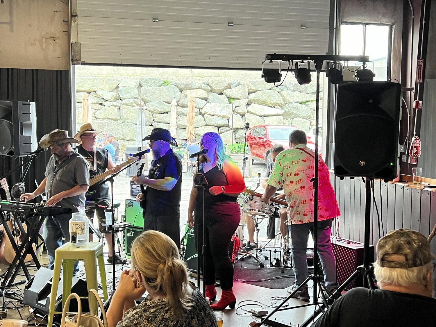 A band performing live at an indoor venue with four musicians. One plays keyboard, two sing into microphones, and the fourth plays guitar. Audience members are seated at tables, with some facing the stage and others outside near a stone wall and park