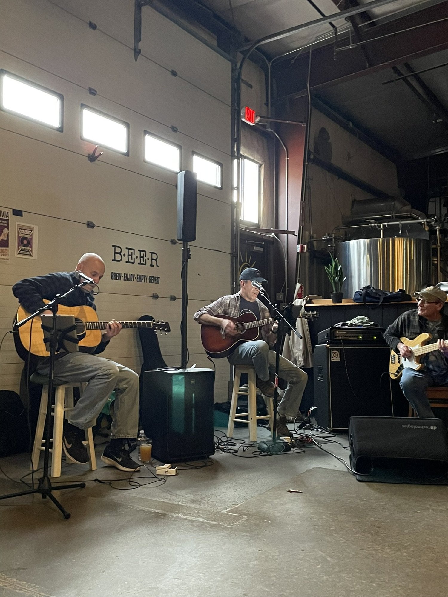 Three musicians performing with acoustic guitars in an indoor venue. There is a wall with the word 'BEER' and the phrase 'brew, enjoy, drink, drink' written on it.