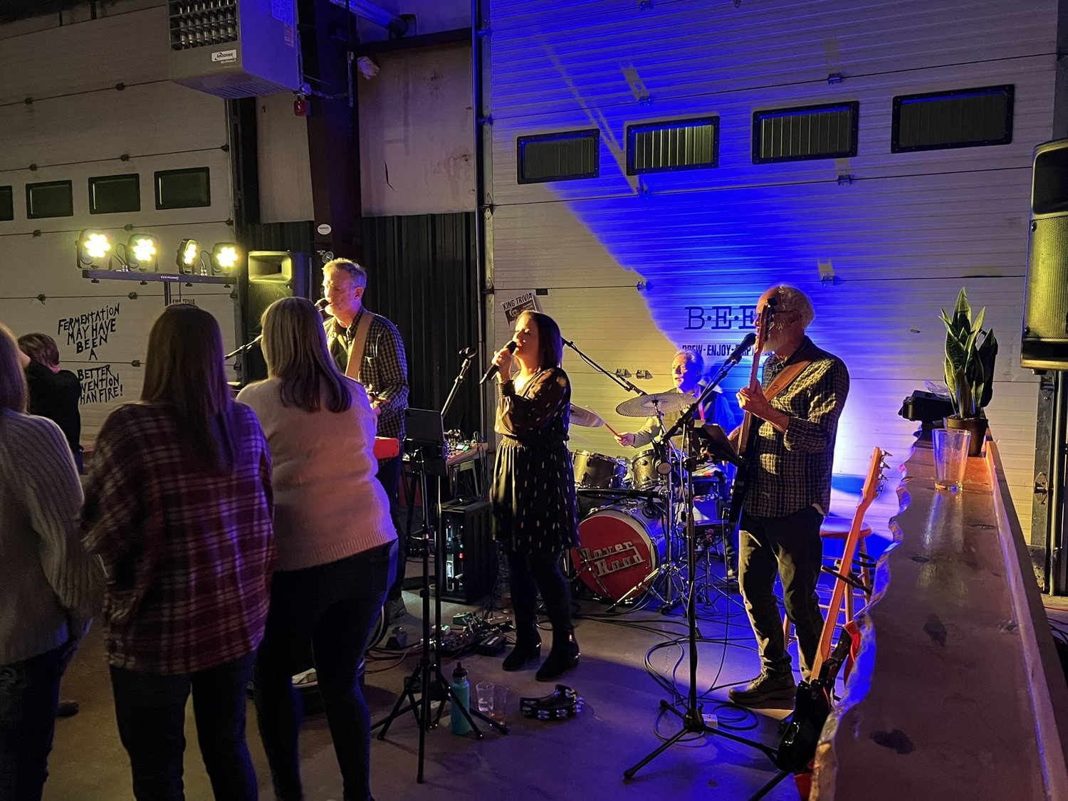 A live band performing on stage with four musicians playing instruments and a female singer. Audience members are watching and standing nearby in a dimly lit setting with blue and yellow stage lights.