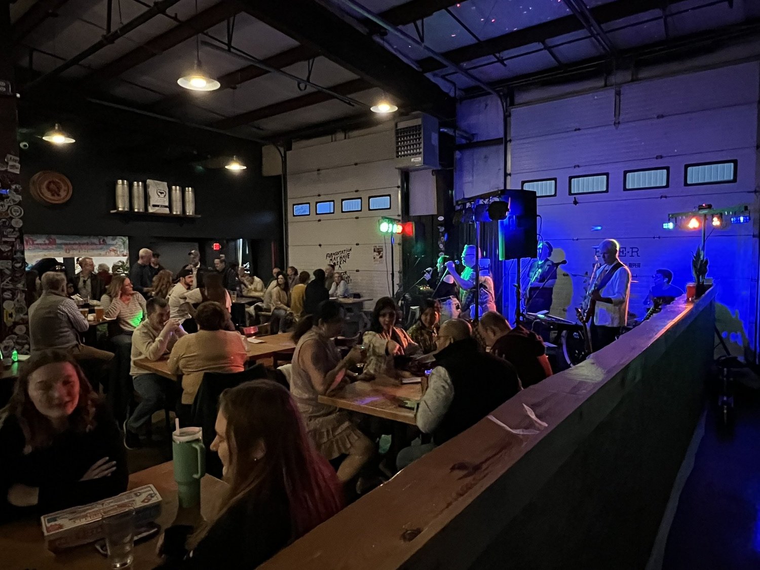 People dining at tables in a bar or restaurant with a live band performing on stage, illuminated by colorful stage lights.