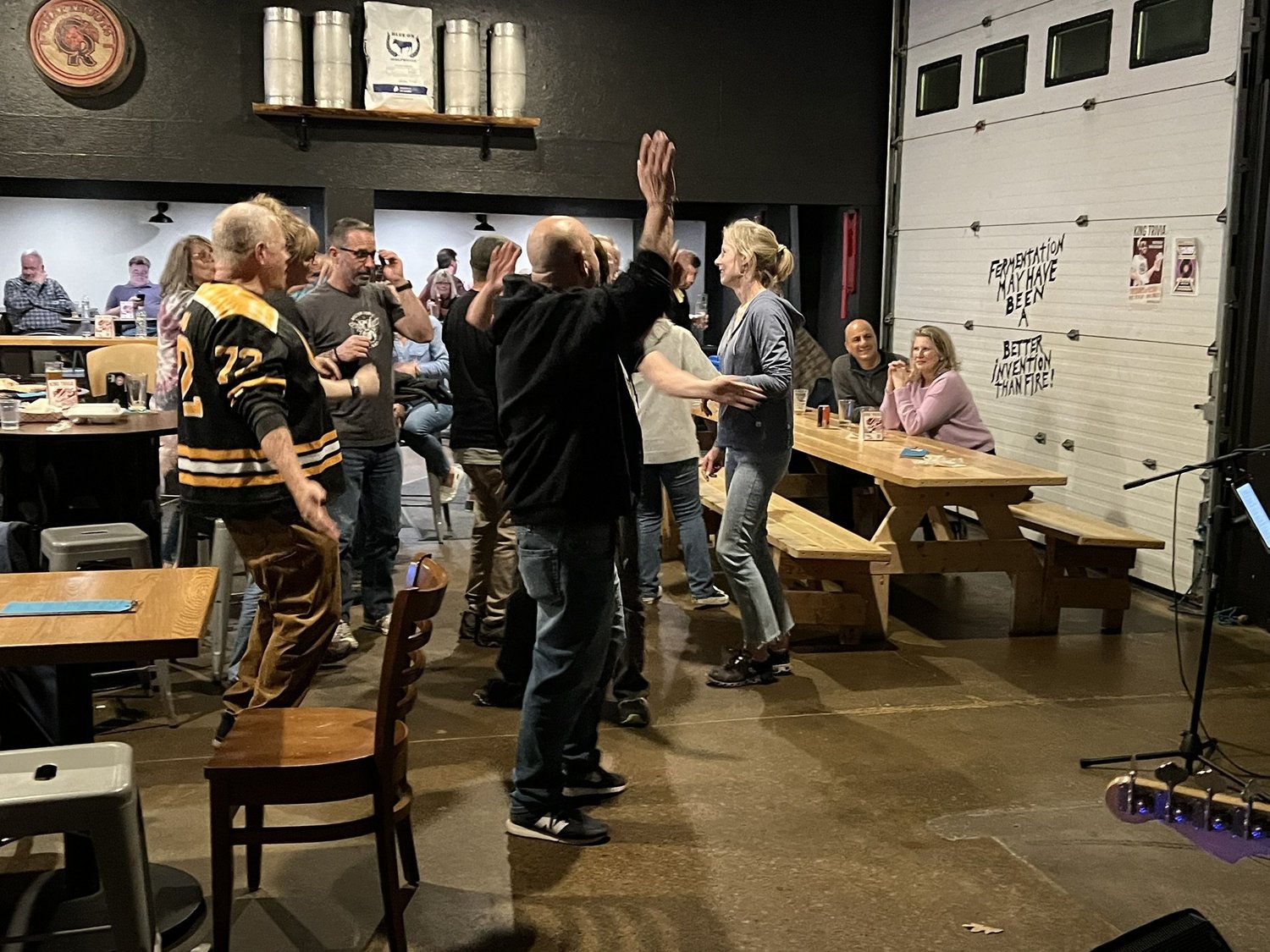 People dancing and socializing in a brewery or bar setting with casual attire. Some are sitting at tables, others are standing, with a wall featuring equipment and humorous text in the background.