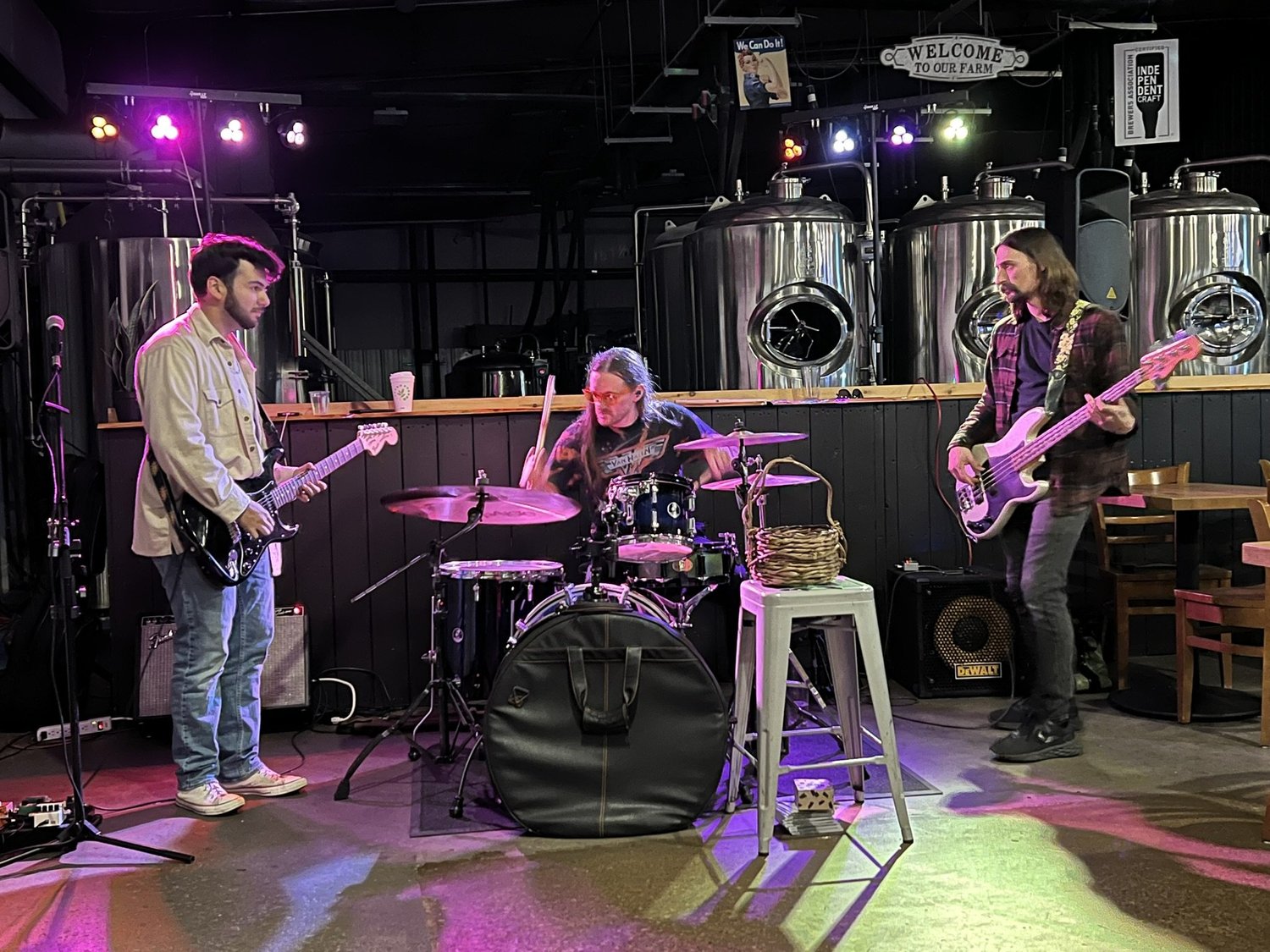 Three musicians performing on stage with instruments: electric guitar, drums, and bass guitar, in a brewery setting with brewing tanks in the background.