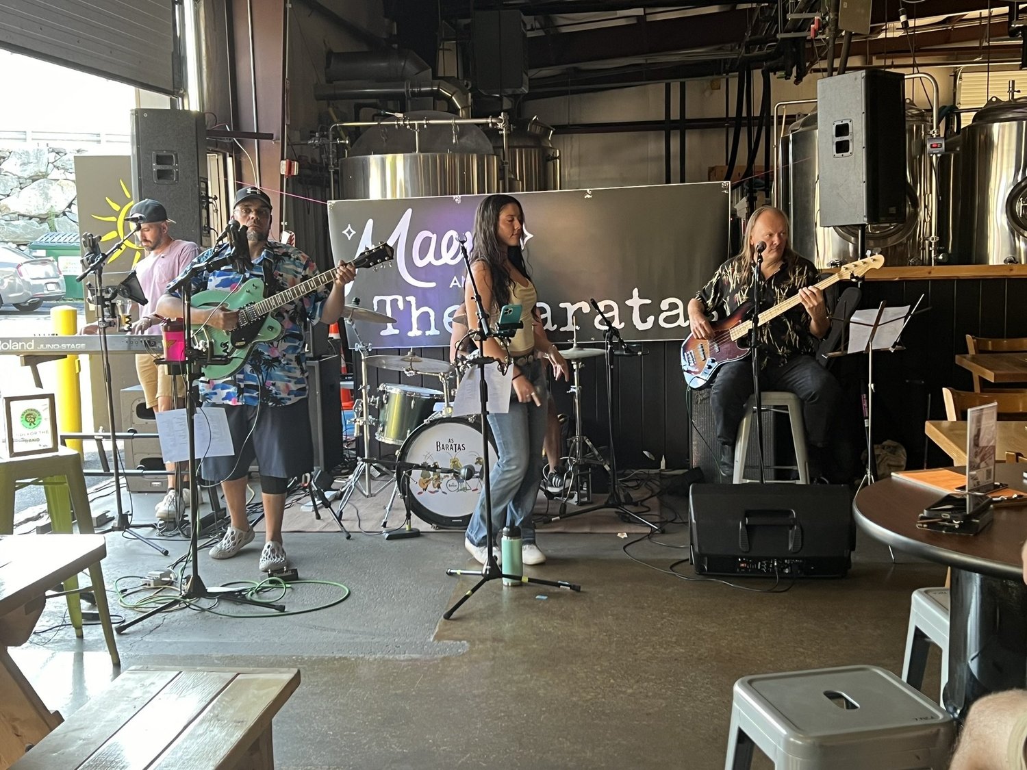 A four-member band performing live in a brewery with brewing tanks in the background. The band includes a drummer, a guitarist, a bassist, and a female singer. There are microphones, music stands, and electronic equipment on stage.