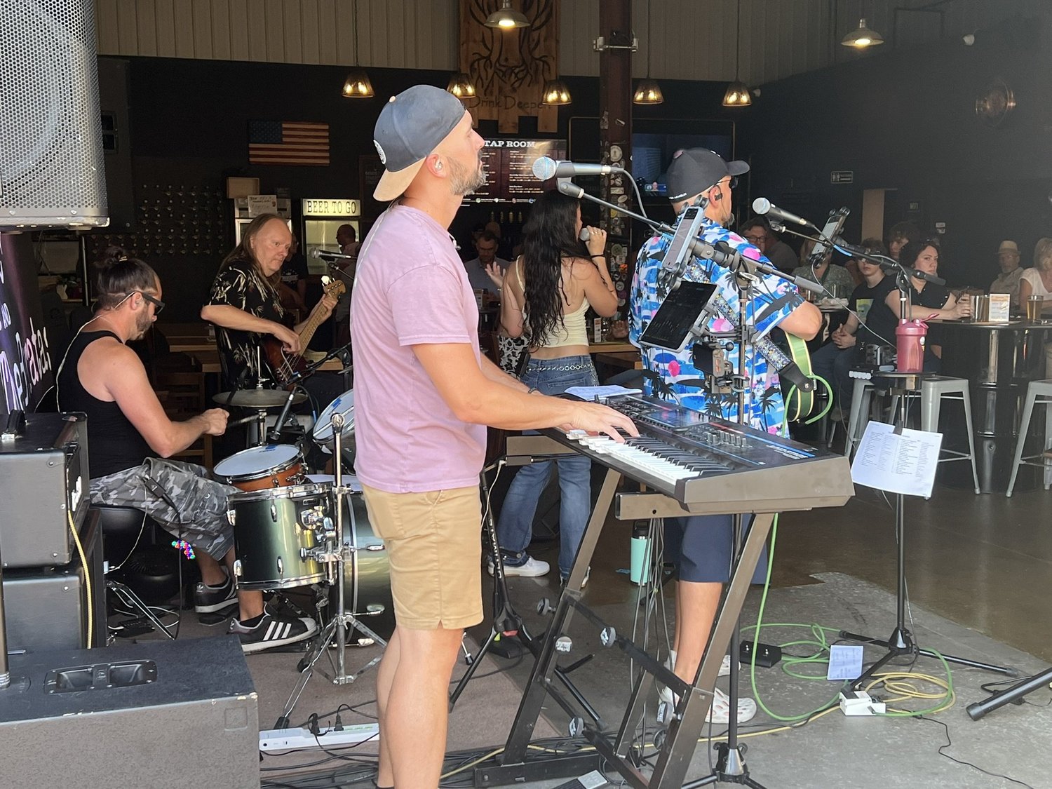 A band performing live at a bar or club, with musicians playing drums, bass, keyboard, and a singer, and an audience watching.