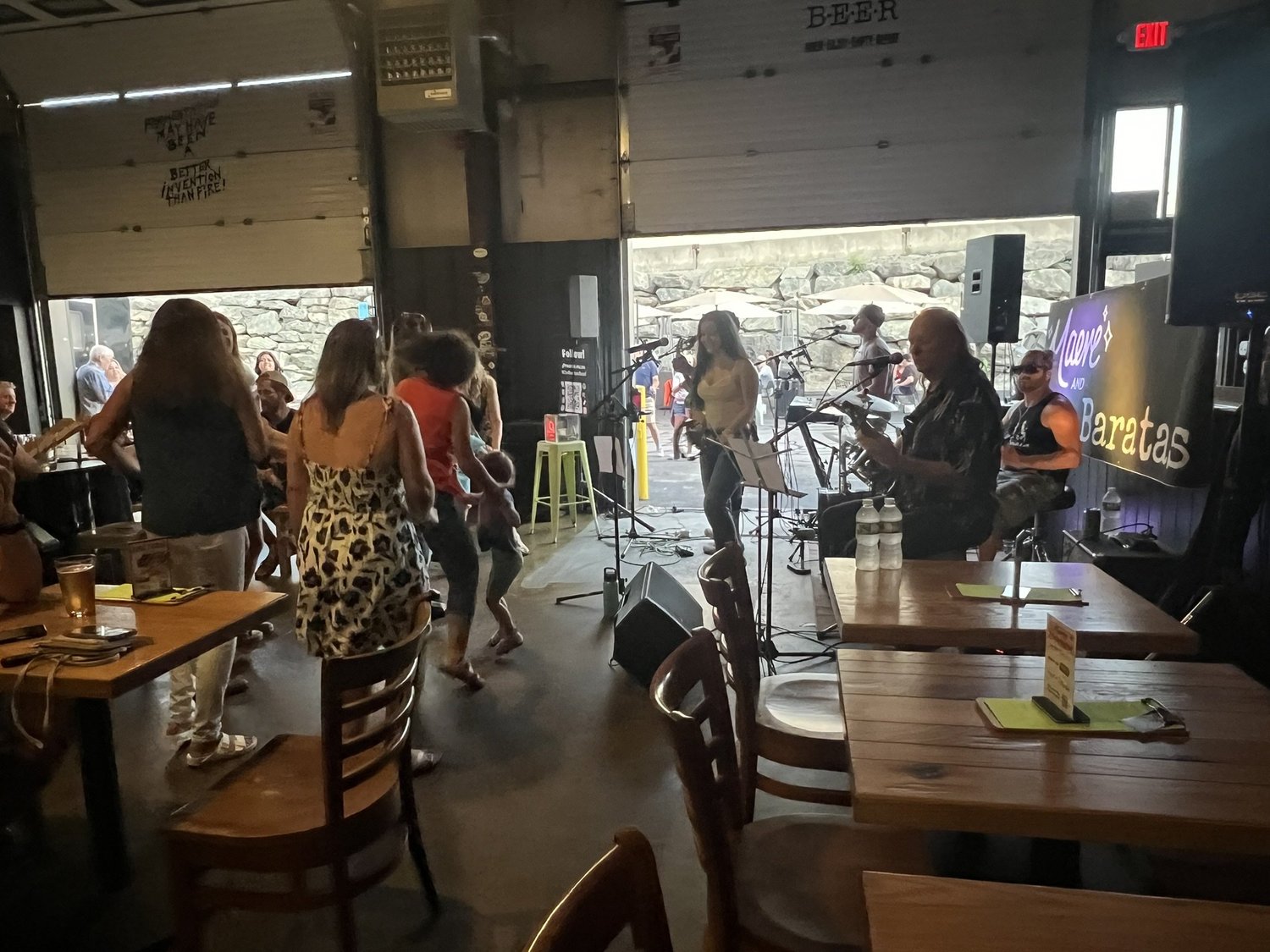People dancing and enjoying live music at a bar or restaurant with tables and chairs, a band performing on stage, and large open doors showing an outdoor area.