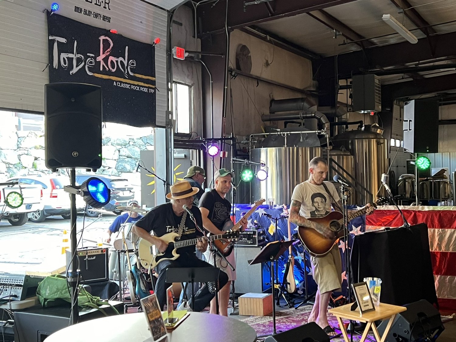 A band performs on stage inside a brewery with stainless steel brewing tanks in the background. The band has three members: one with a hat playing guitar and singing into a microphone, another playing guitar, and the third playing a bass guitar. Colo