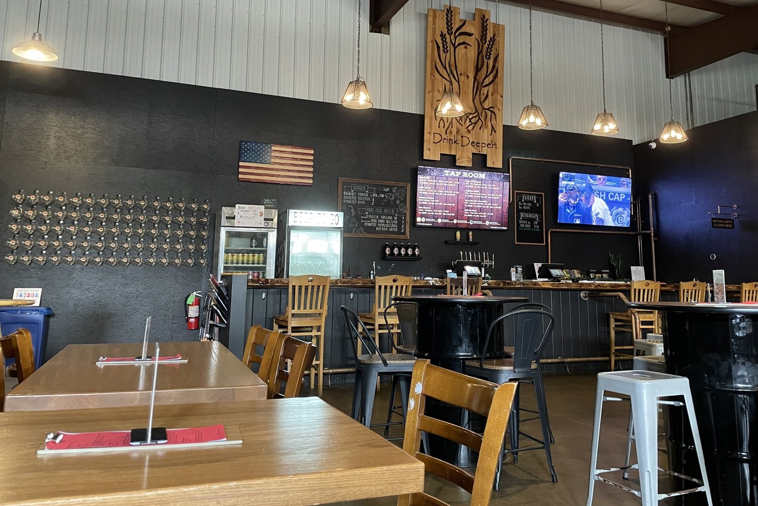 Interior of a restaurant or bar with wooden tables and chairs, a chalkboard menu, digital screens, a wall-mounted American flag, and a decorative wooden panel with the words 'Drink Deeper'.